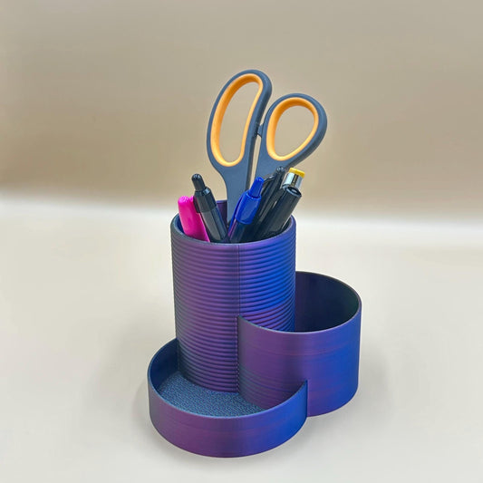 May include: A purple and black desk organizer with a cylindrical compartment for pens and a curved compartment for other items. The organizer contains a pair of black scissors, a pink pen, a blue pen, and a black pen.