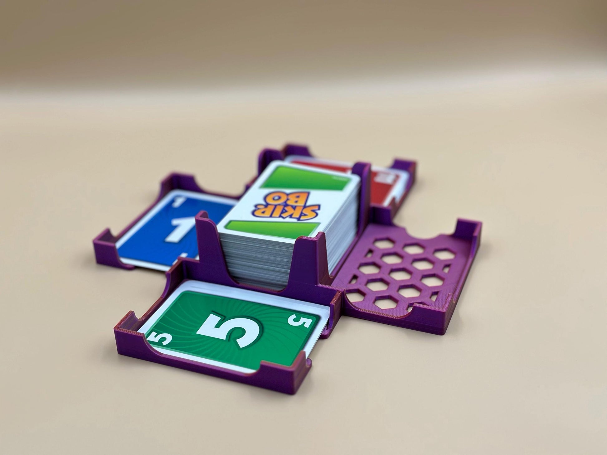 May include: Purple card holder with four slots for cards. The card holder is holding a deck of cards with the words &#39;Skip Bo&#39; on the top card and a card with the number &#39;5&#39; on it. The card holder has a honeycomb pattern on the bottom.